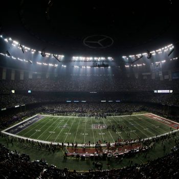 Stadium crowd watches football game; bright floodlights illuminate the field surrounded by fans in large indoor arena.
