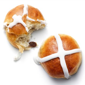 Two glazed buns, one partially eaten, reveal raisins inside; both display white icing crosses on a plain white background.