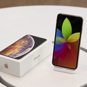 Smartphone displays colorful flower pattern, standing on a white dock beside its box, on a circular white table.