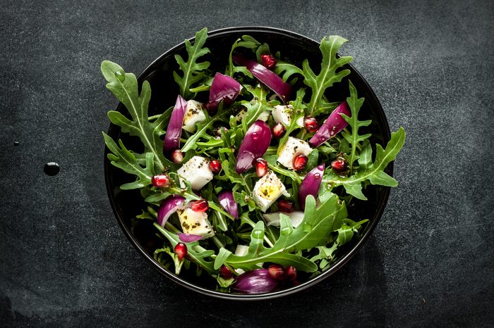 Fresh spring salad with rucola, feta cheese, red onion and pomegranate seeds in black bowl on chalkboard background.