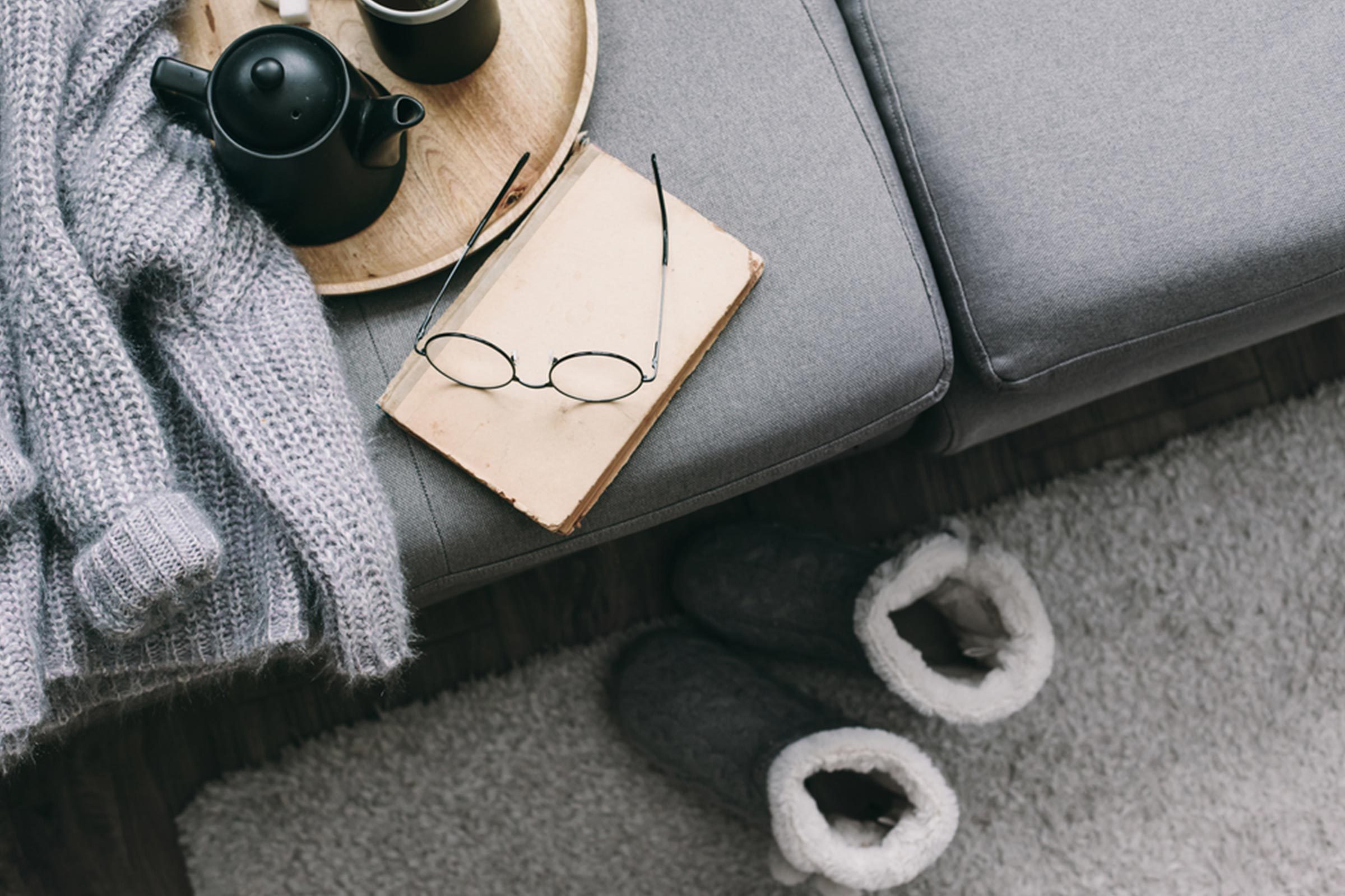 Glasses rest on a closed book beside a cozy sweater, with a teapot on a tray atop a gray couch in a relaxed setting.