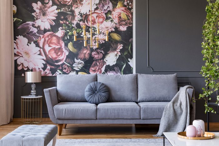 Blanket on grey couch in living room interior with flowers wallpaper and lamp on table.