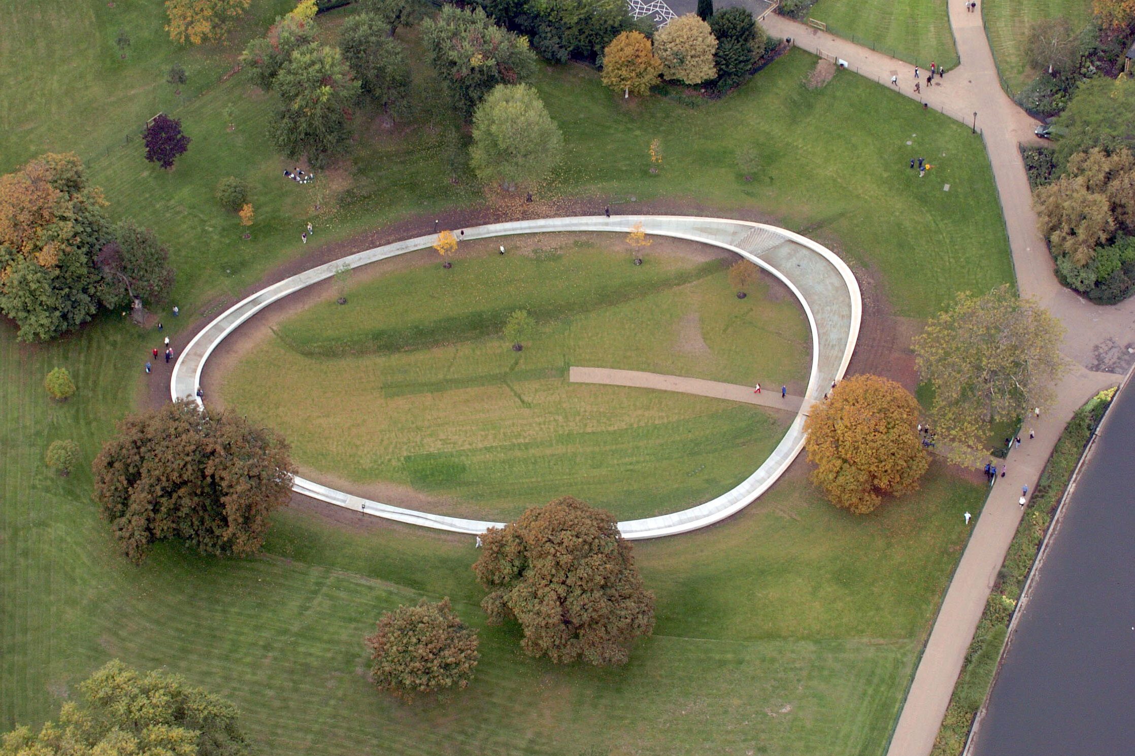 Oval water feature meandering through park surrounded by trees and pathways, people strolling leisurely, creating a serene atmosphere.