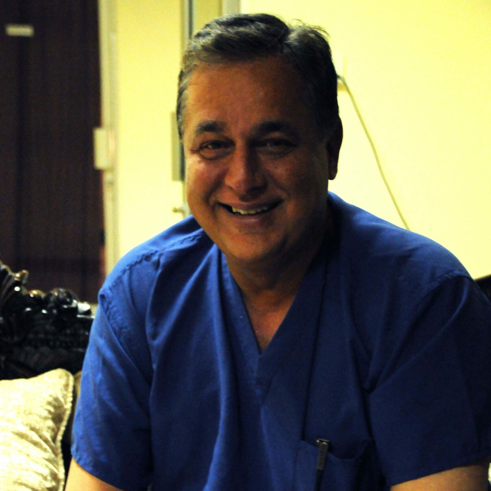 A person is smiling while sitting indoors, wearing blue scrubs, with a neutral background featuring a door and a sofa with cushions.