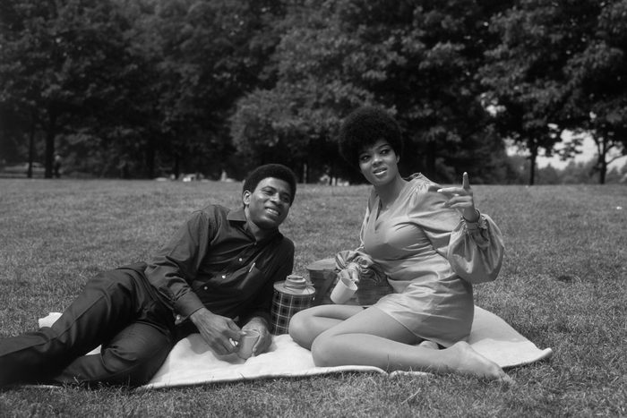 Two people relax on a picnic blanket, enjoying drinks, in a grassy park surrounded by trees.