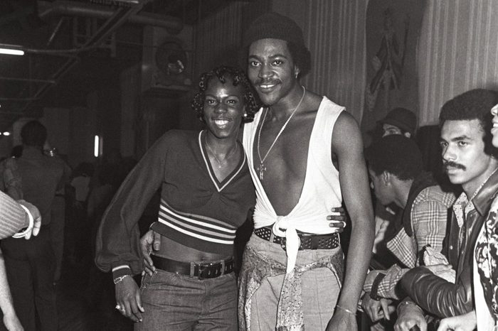 Two people pose on roller skates in a busy indoor rink, surrounded by onlookers against a background of patterned walls and ceiling beams.