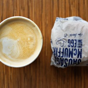 A cup of cappuccino sits beside a wrapped breakfast sandwich on a wooden table.