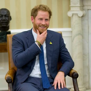 Man smiles, touching chin, seated in an elegant room with columns and a bust in the background. Wearing a suit, bracelets, and blue tie.