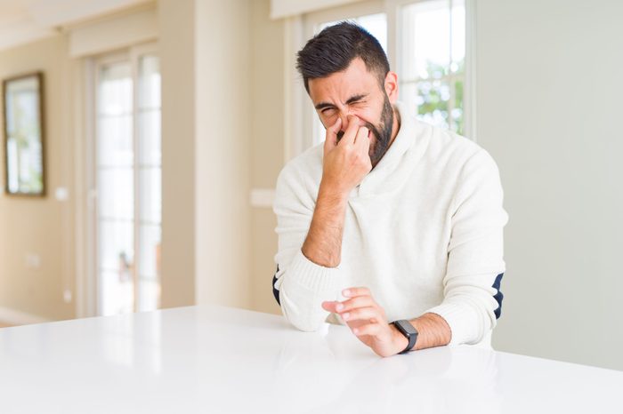 Handsome hispanic man wearing casual white sweater at home smelling something stinky and disgusting, intolerable smell, holding breath with fingers on nose. Bad smells concept.