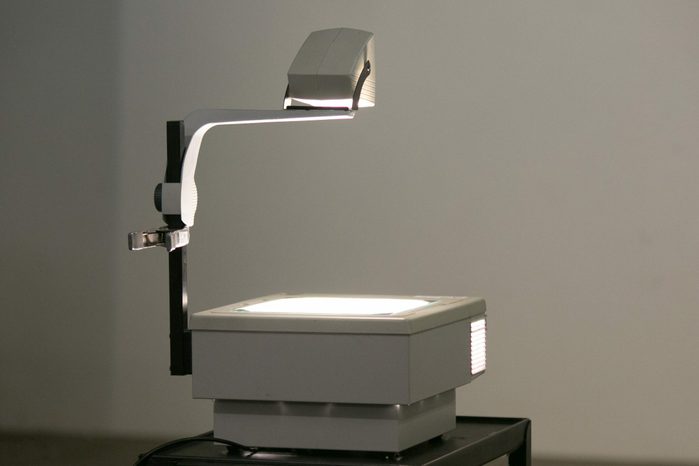 An overhead projector illuminates, casting light onto a blank wall, in a dimly lit room, with cables trailing onto a small table.