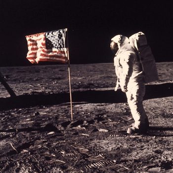 Astronaut stands beside a planted American flag on the moon's surface, with spacecraft landing legs visible to the left.