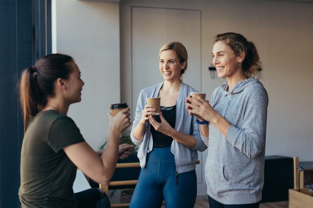 Three women hold coffee cups, smiling and chatting in a bright indoor setting.