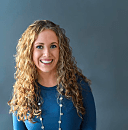 Smiling woman with curly hair wears a blue top and necklace, seated against a plain gray background.