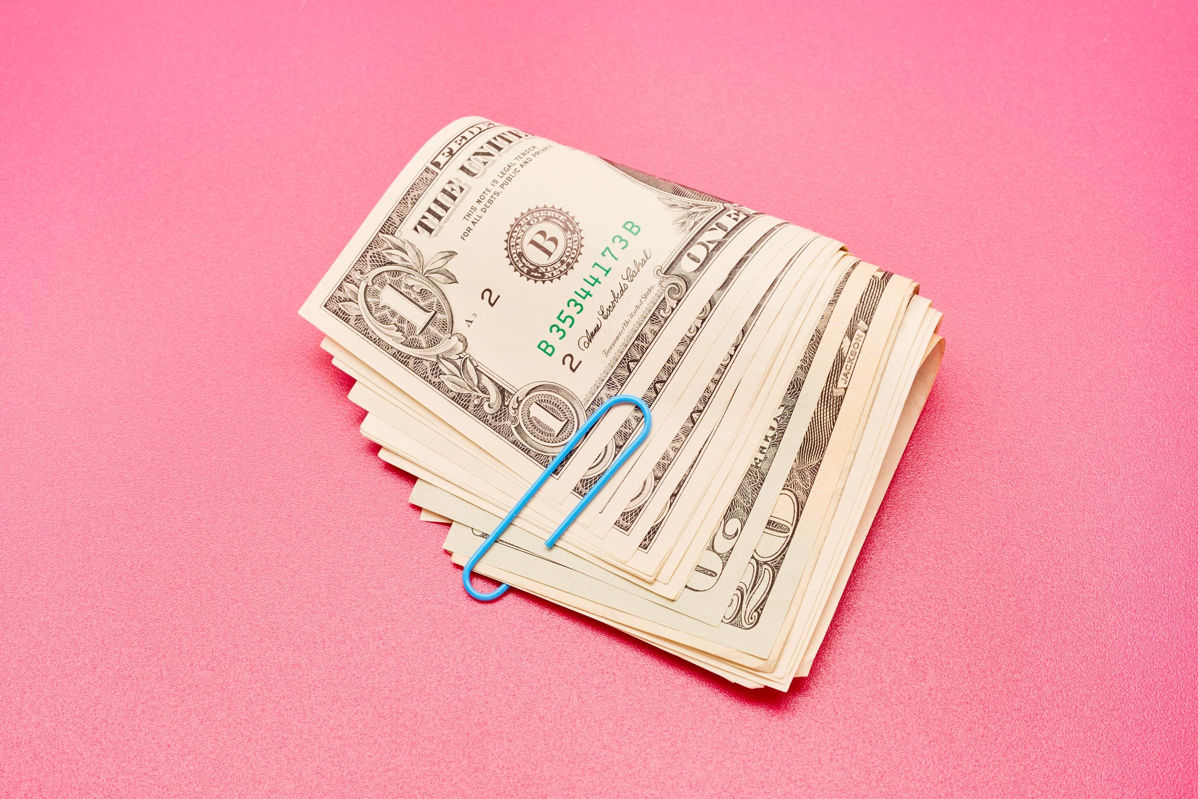A stack of one-dollar bills secured by a blue paperclip rests on a smooth pink surface.