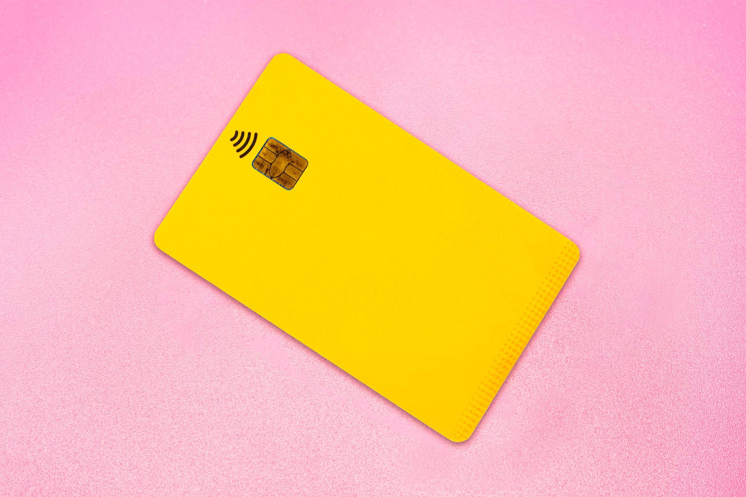 A yellow credit card lies on a pink surface, displaying a chip and contactless symbol.
