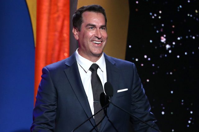 A man in a suit smiles while speaking at a podium, with a colorful curtain and starry backdrop behind him.
