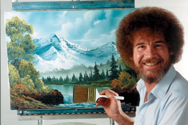 Artist smiles while holding a paintbrush, painting a scenic landscape with mountains and trees on an easel. Bright studio setting.