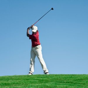 Golfer swings club, poised mid-swing, on a grassy hill with a clear blue sky background.