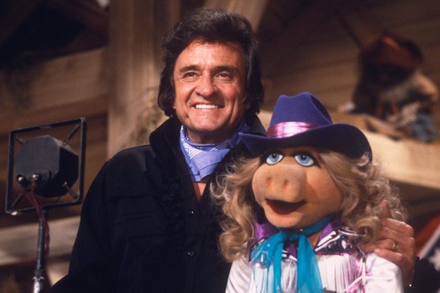 Man and puppet smile together indoors, with the puppet wearing a cowboy hat. A microphone is visible to the left.