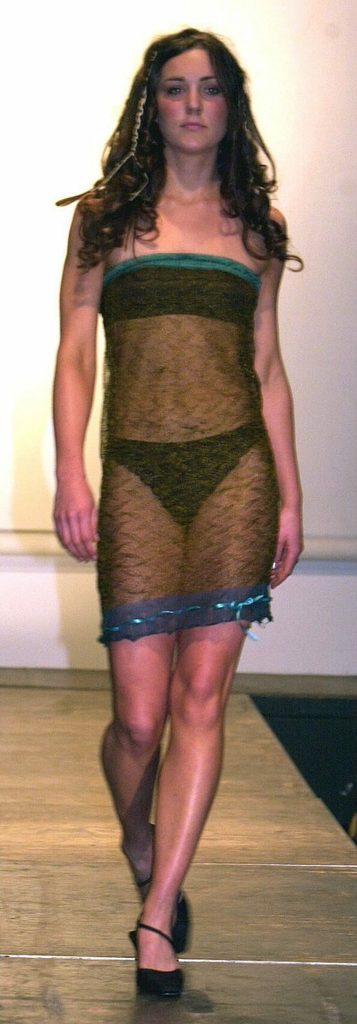 Model wearing a sheer dress walks confidently on a runway in a fashion show setting.