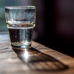 Glass half-filled with water, casting a shadow on a sunlit wooden table in a softly lit room.