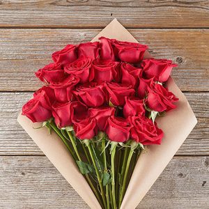 A bouquet of red roses rests on brown paper, lying on a rustic wooden surface.