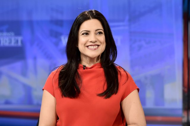 A woman in a red dress smiles confidently; she is seated in a studio setting with a blue-toned backdrop.