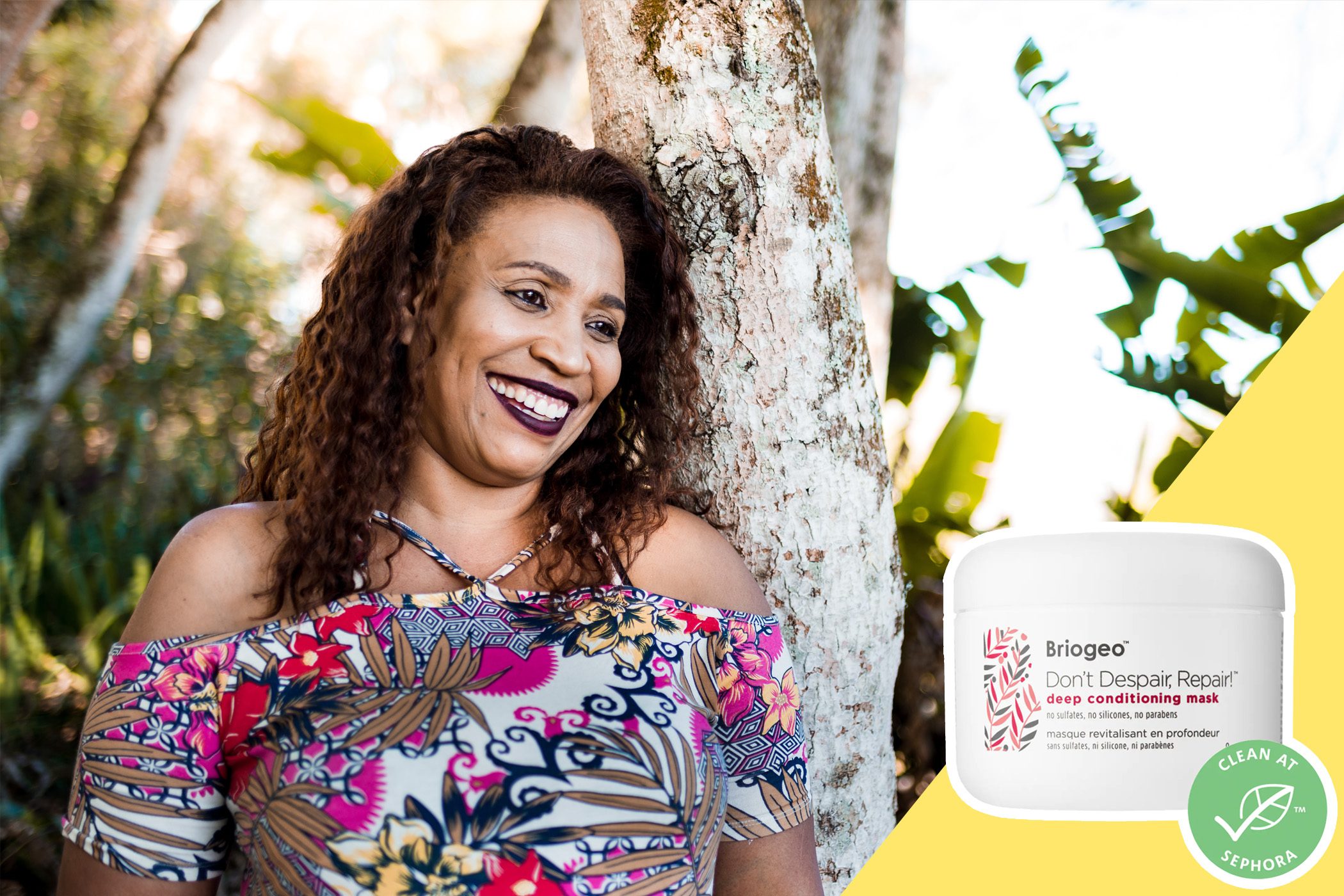 Container rests beside smiling woman leaning on tree, wearing colorful floral dress, in sunny outdoor setting. Text: "Briogeo Don't Despair, Repair! deep conditioning mask."