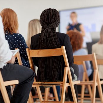 Audience faces presenter in a seminar room, seated on wooden chairs, focusing on a screen.