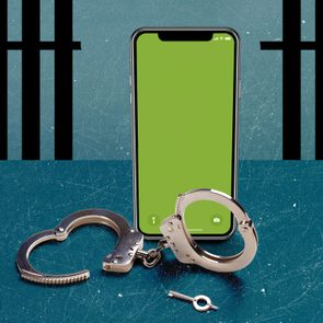 Smartphone stands upright beside open handcuffs and a key on a scratched blue surface, with black prison bars in the background.