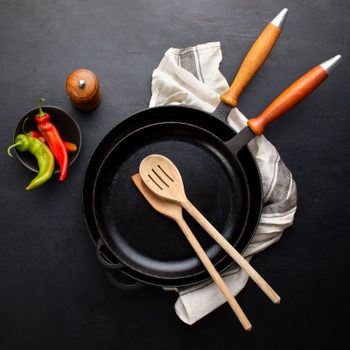 Skillet holds wooden spoons, resting on a cloth; adjacent, a bowl of peppers and a pepper mill add color against a dark surface.