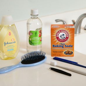 Hairbrush, toothbrush, and tweezer rest on a bathroom counter alongside shampoo, vinegar, and baking soda near a faucet.
