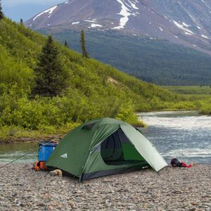 A green tent stands on a rocky riverside, surrounded by mountains and greenery, with nearby camping gear.
