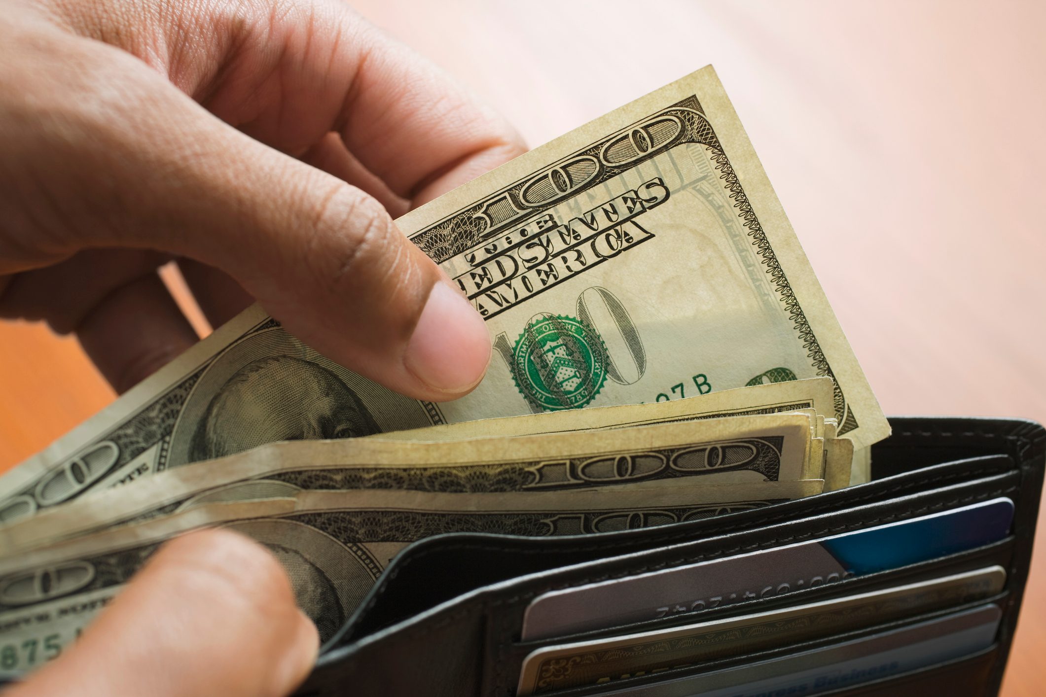 Hand withdrawing hundred-dollar bills from a black wallet containing several credit cards, set against a blurred background.