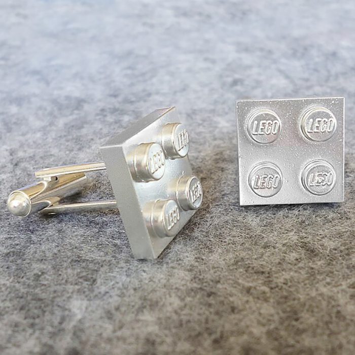 Silver cufflinks resemble LEGO bricks, lying on a gray felt surface. Each has 