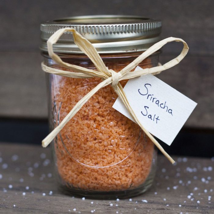 A jar contains orange Sriracha salt, tied with raffia and labeled, placed on a rustic wooden surface.
