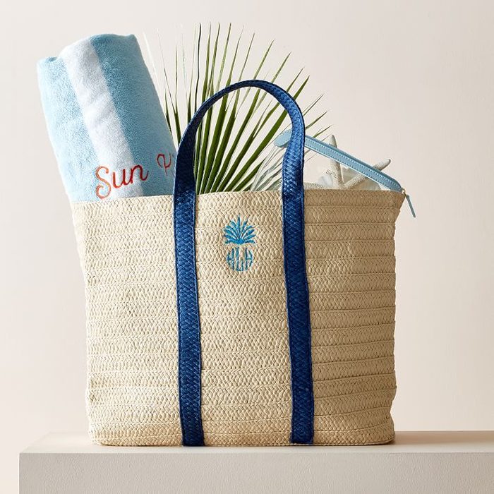 A straw tote bag with blue handles holds a beach towel, palm frond, and accessories on a white platform.