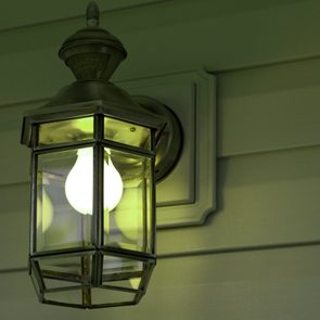 A wall-mounted outdoor lantern illuminates with a bright bulb, surrounded by green wooden siding.