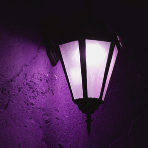 Lantern emits soft purple light, illuminating a textured wall in a dim, dark setting.
