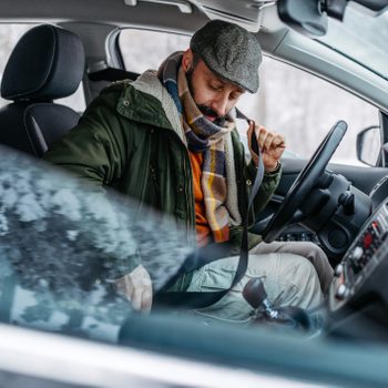 Man fastening seatbelt, wearing a cap and scarf inside a parked car, surrounded by a snowy outdoor environment.