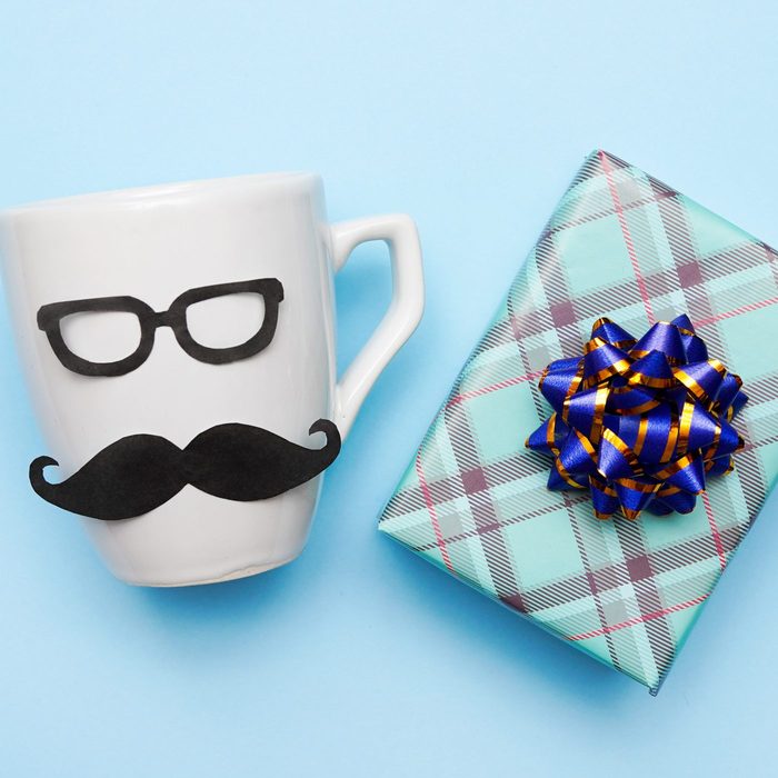 White mug with black glasses and mustache alongside a gift-wrapped box, blue plaid paper, and navy-gold ribbon, set on a light blue background.
