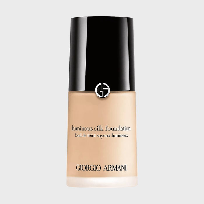 Foundation bottle displays Giorgio Armani branding, featuring black cap and beige base, set against a plain, light gray background. Text: 