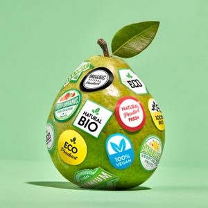 A pear covered in stickers promoting organic, vegan, and eco-friendly qualities sits on a green background.