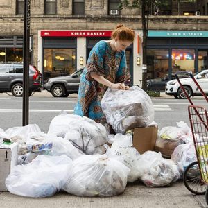 Person examines trash bags on sidewalk; stores in background and cars on street.