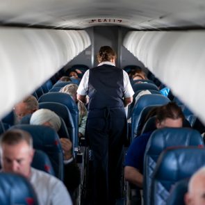 Flight attendant walks down the aisle inside a crowded airplane, passengers seated, overhead compartments closed.