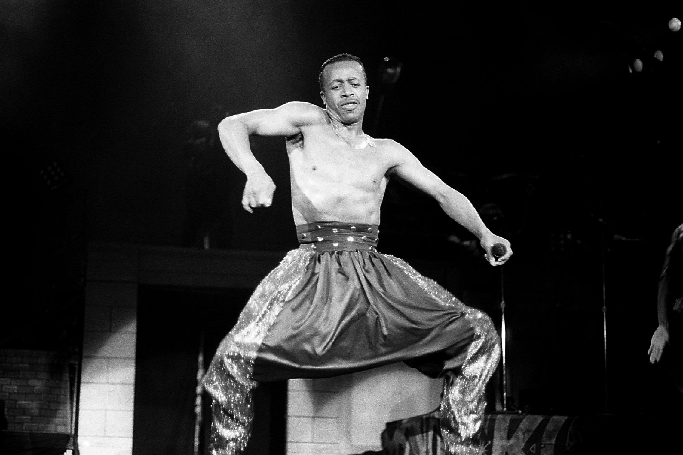 A shirtless man energetically dances on stage, holding a microphone, wearing shiny, loose pants in a dark performance setting.