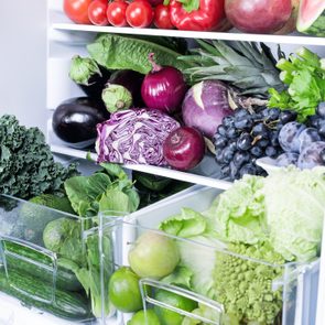 A refrigerator holds assorted fruits and vegetables, like tomatoes, red cabbage, lettuce, eggplants, grapes, and apples, neatly arranged on shelves.