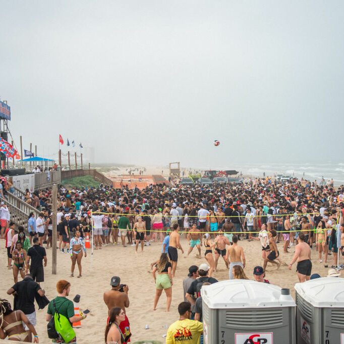 Crowd gathers excitedly, playing beach volleyball, surrounding a stage at a lively seaside event with portable toilets and flags in the background.
