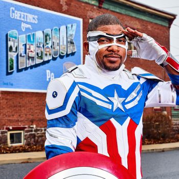 Person salutes wearing superhero costume with star emblem, standing in front of a brick wall with "Greetings from PENBROOK Pa" mural.