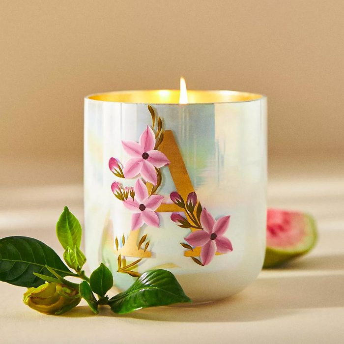 Candle burns brightly, decorated with pink flowers, on a neutral surface beside green leaves and a slice of fruit.